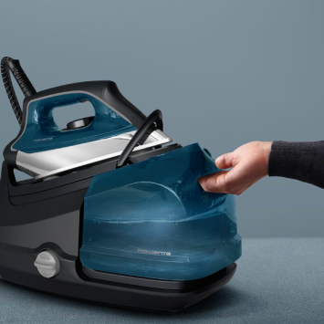 A hand interacts with a sleek, dark blue steam station. The iron rests on a transparent base, revealing the water level. Set on a dark gray surface with a light blue background.